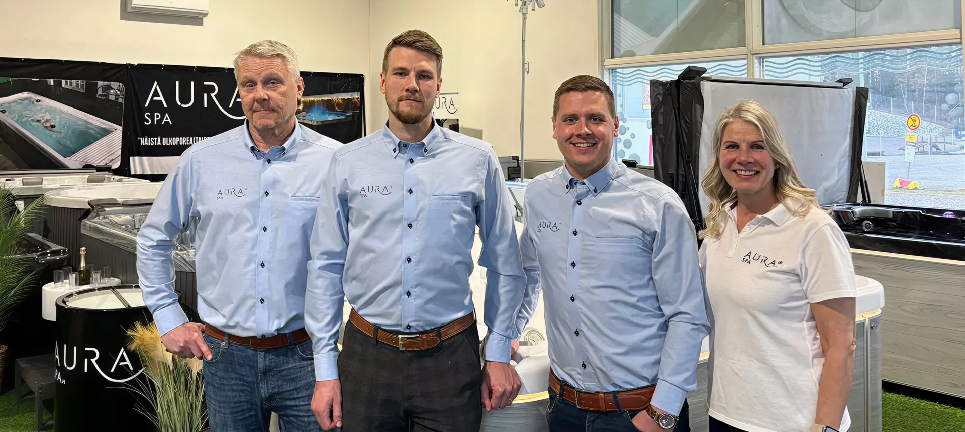 Four Aura Spa staff posing together at a showroom booth, smiling in light blue shirts and a white polo shirt.