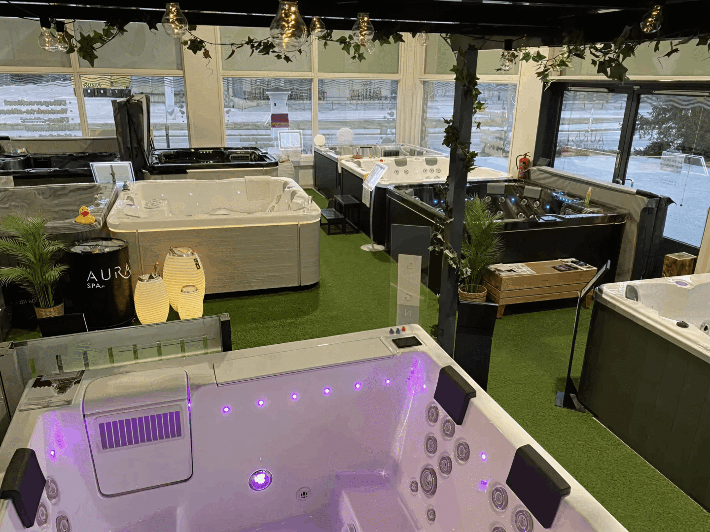 Indoor showroom displaying multiple hot tubs and spas on artificial grass flooring, with decorative hanging lights and plants.