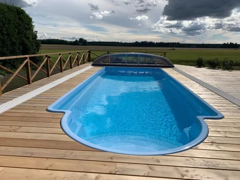 Outdoor pool on a wooden deck with a curved front edge, clear blue water, and a glass cover at the far end, open countryside in the background