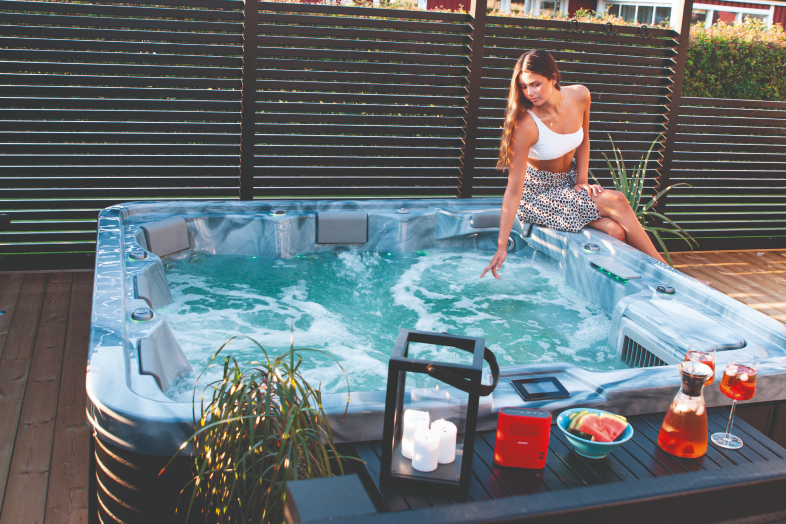 Woman in a white crop top and leopard-print skirt sits on the edge of a bubbling hot tub on a wooden deck, touching the water.