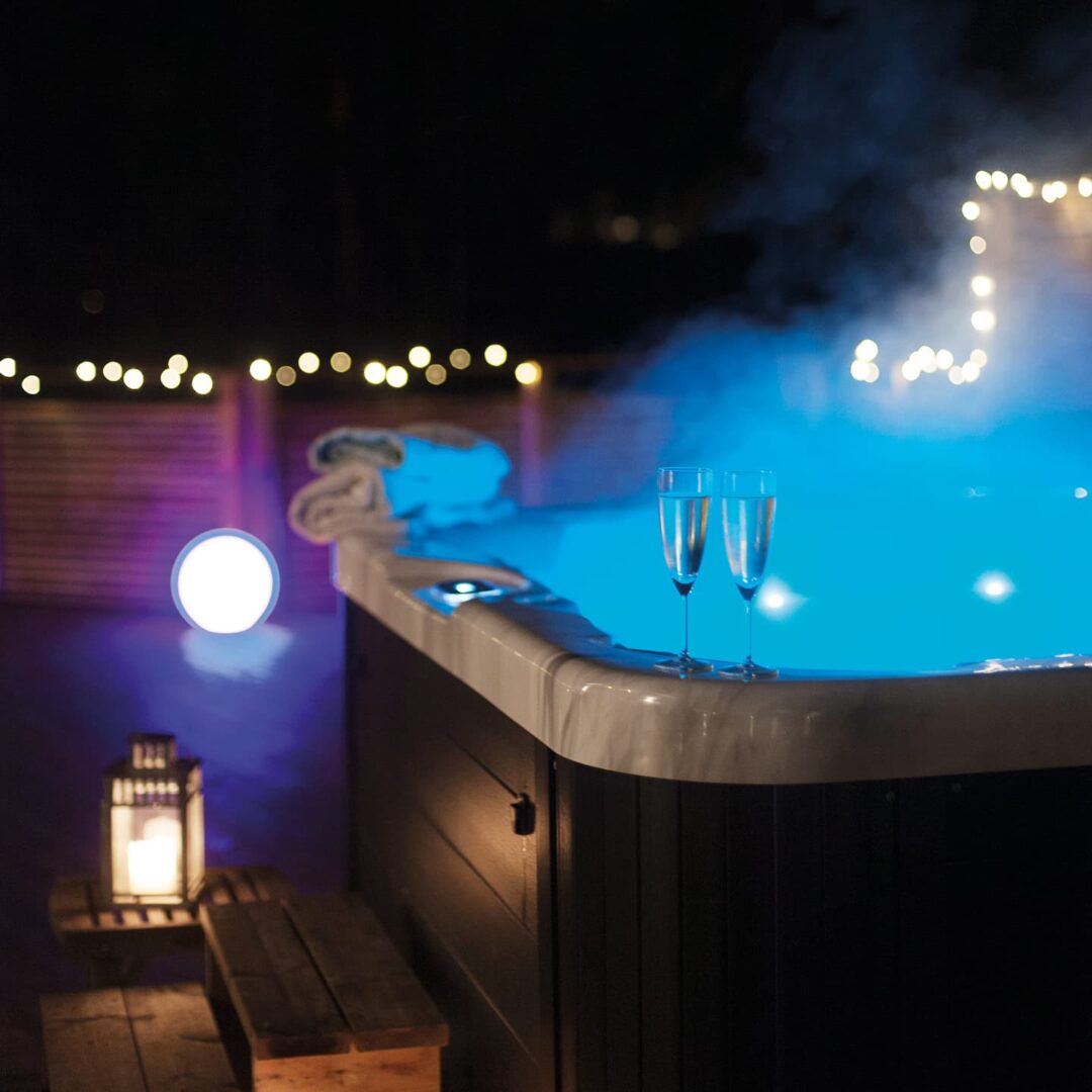 Nighttime hot tub scene with two champagne flutes on the edge and soft blue water glow, string lights in the background, lantern on a wooden step