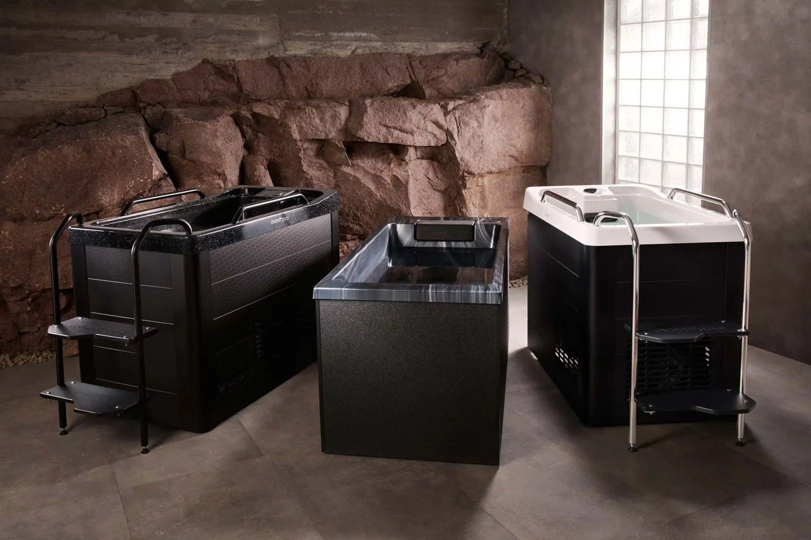 Indoor showroom displaying three luxury hot tubs with dark exteriors, rock wall backdrop, and a glass-block window to the right